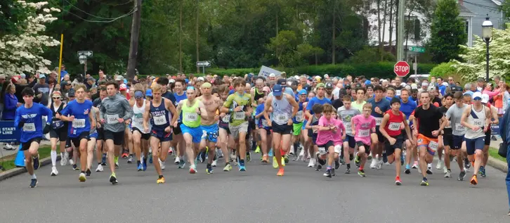 A large field of runners of all ages surging from the Doylestown 5K start line on a tree-lined residential street on a spring morning